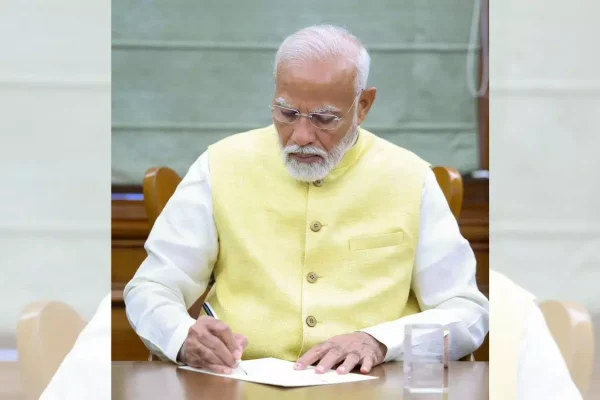 పసుపు రంగు జాకెట్ ధరించిన ప్రధాని నరేంద్ర మోదీ టేబుల్ వద్ద పత్రంపై సంతకం చేస్తున్నారు
