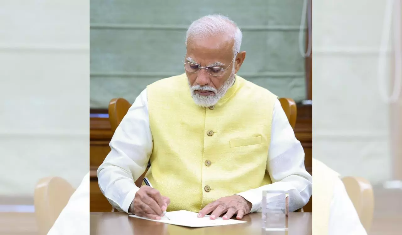పసుపు రంగు జాకెట్ ధరించిన ప్రధాని నరేంద్ర మోదీ టేబుల్ వద్ద పత్రంపై సంతకం చేస్తున్నారు